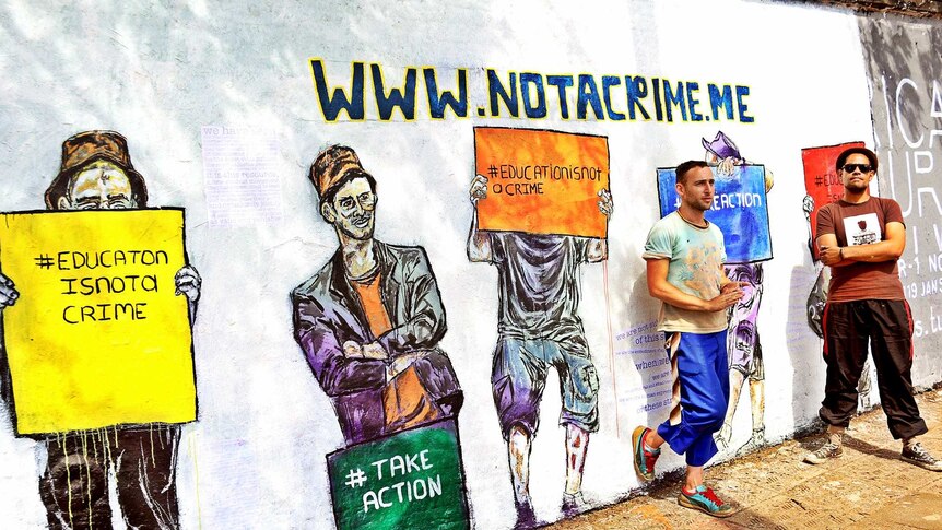 Two artists stand beside their #notacrime mural which shows people holding placards supporting education.