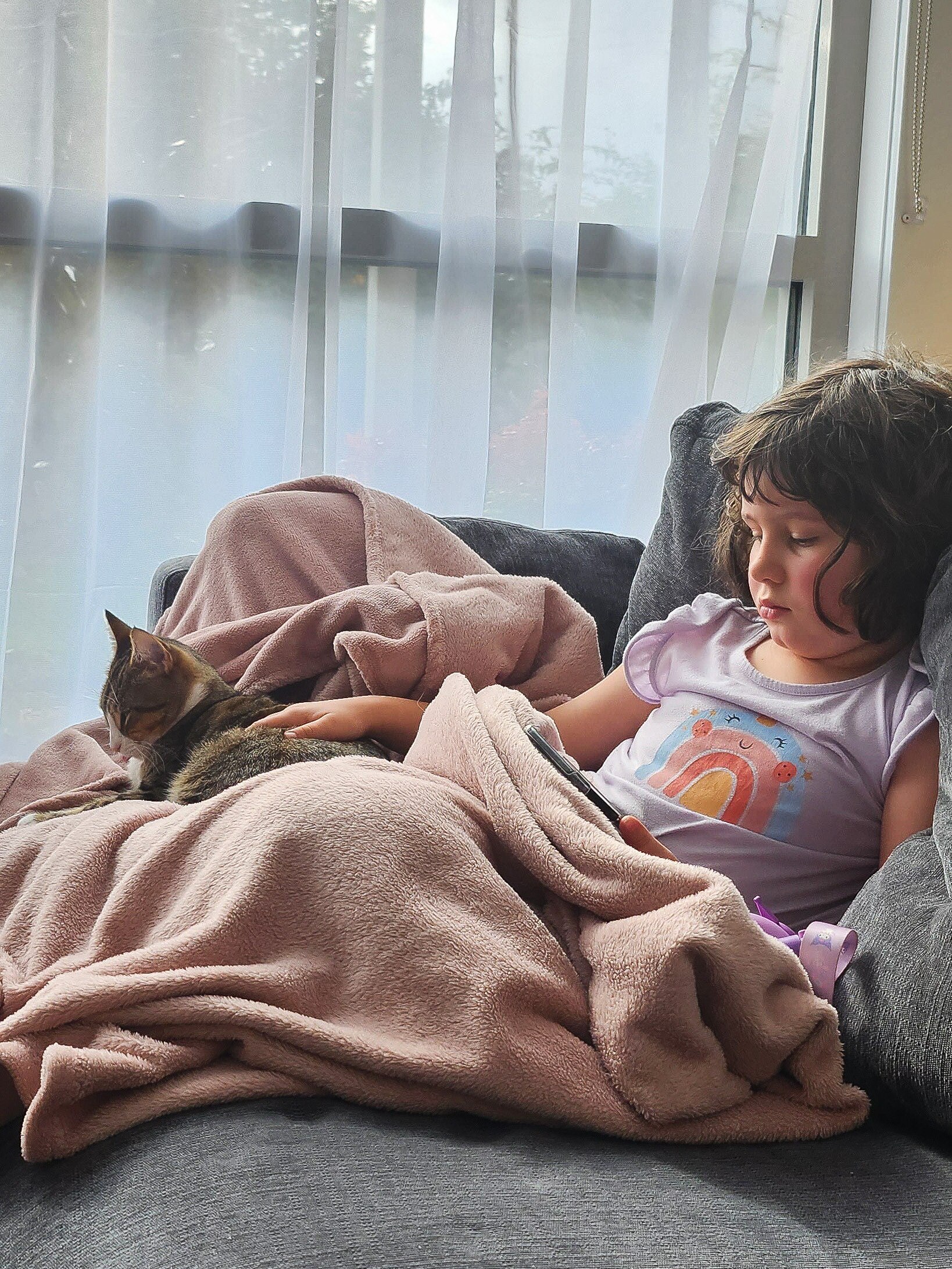 Ada under a blanket on the couch looking miserable, while a cat sits on her lap.