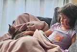 Ada under a blanket on the couch looking miserable, while a cat sits on her lap.