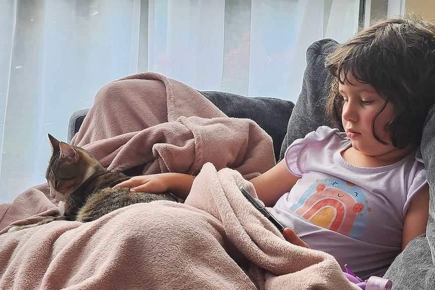 Ada under a blanket on the couch looking miserable, while a cat sits on her lap.