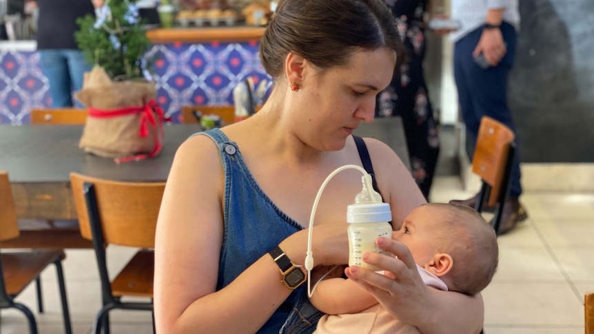 Woman nurses infant in cafe. She also holds a baby bottle filled with milk. A silicon tube is attached to the bottle's teat