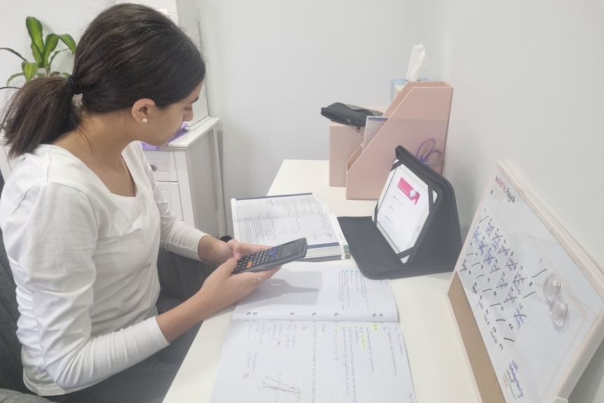 A young girl sits at a desk reading a textbook, calculator in hand. On the wall in front of her is a detailed timetable