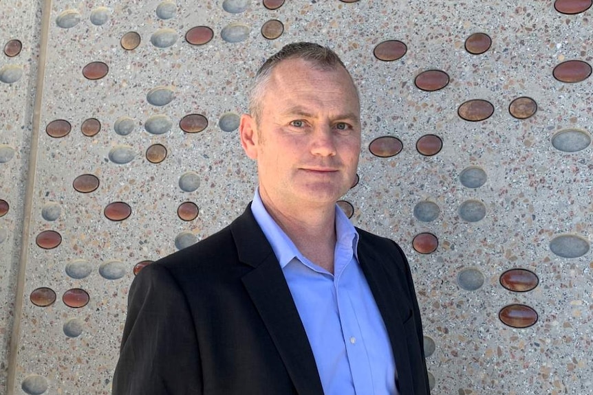 A man in a dark suit jacket, light blue shirt and no tie, stands in front of a wall patterned with spots of different shades