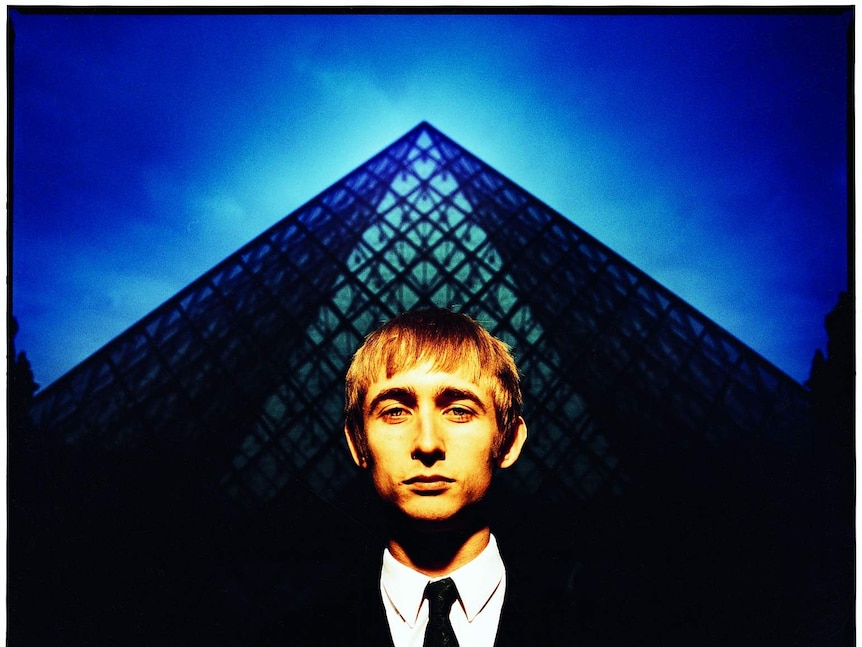 A young besuited man stands in front of a glass pyramid.