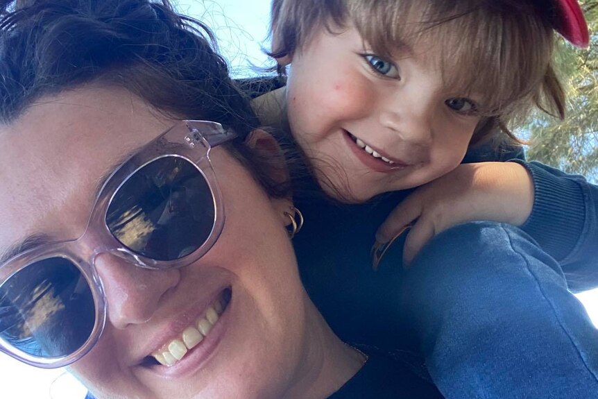 Close up shot of Bridie Jabour in sunglasses smiling with her young child, also smiling, sitting on her shoulders.