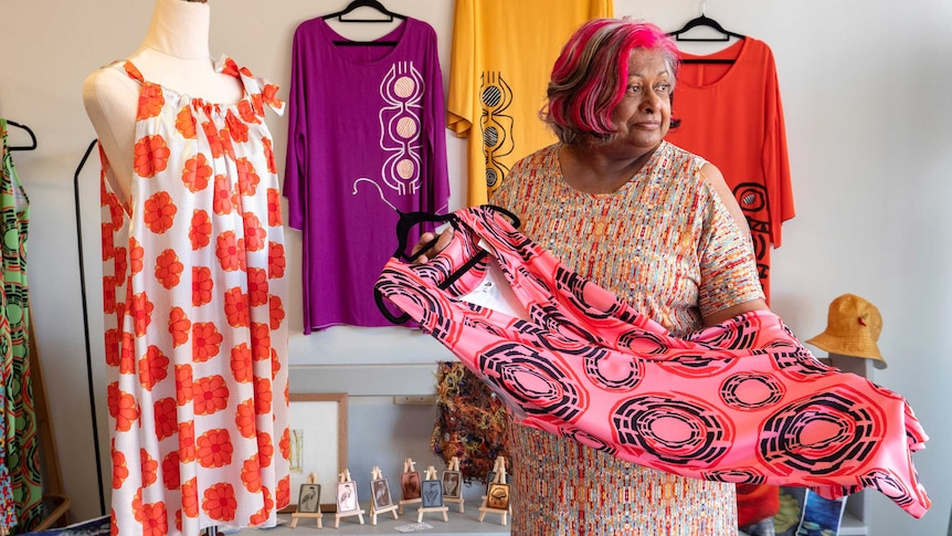 A woman stands holding dresses in a shop setting.