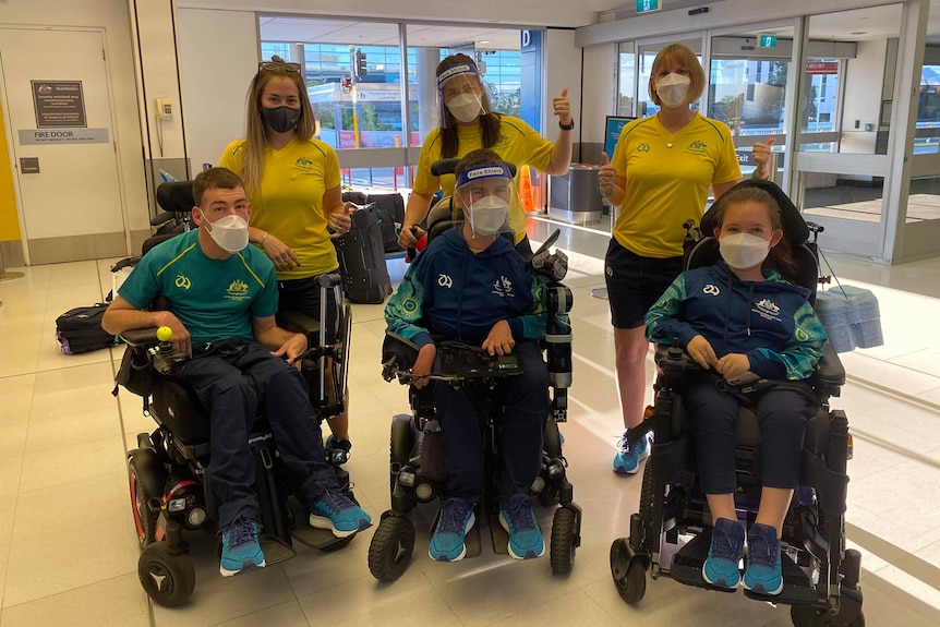 Three women wearing yellow shirts and masks give a thumbs up behind two men and a woman in green shirts in modified wheelchairs.