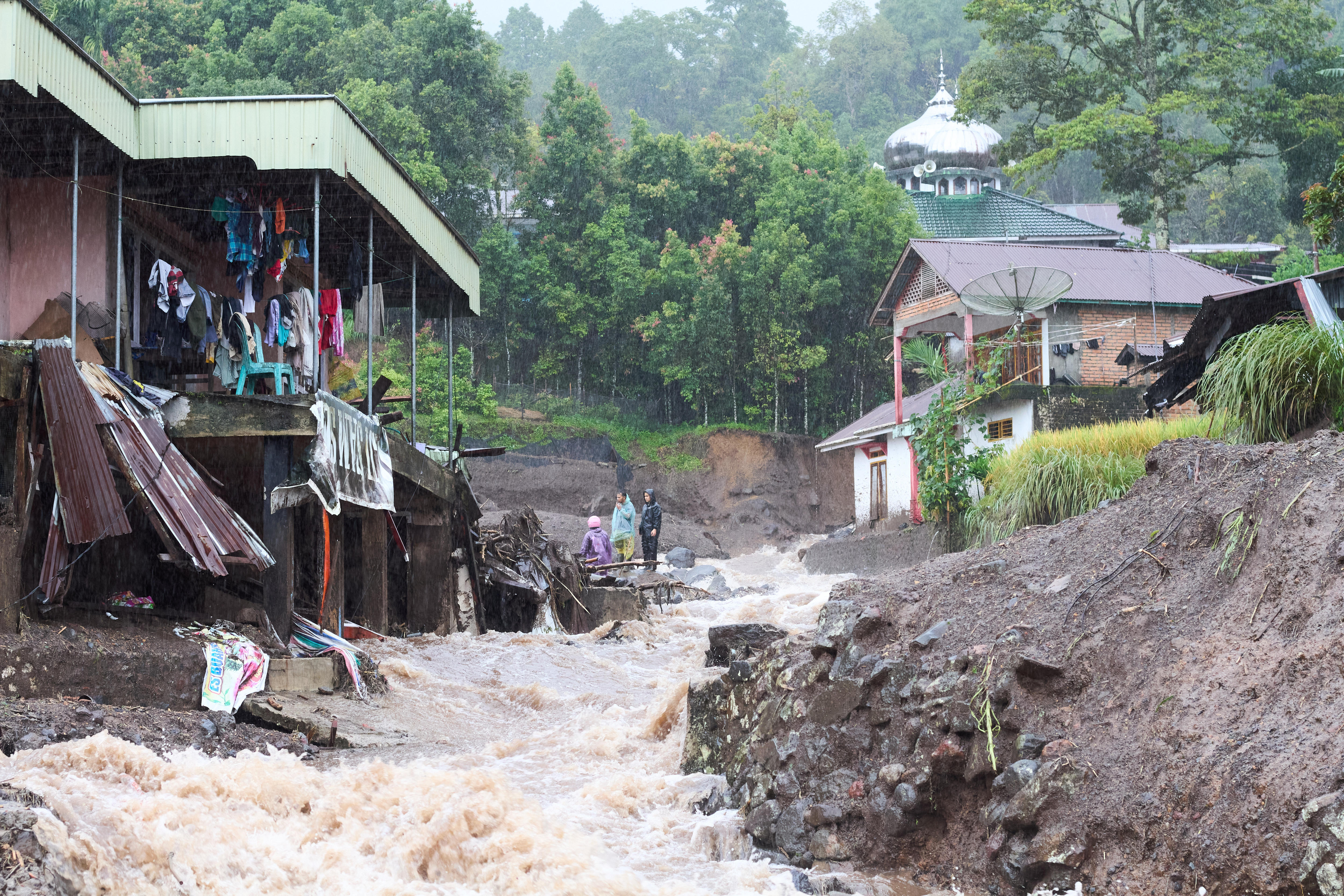 Un río crecido pasa por dos casas, mostrando una severa erosión en el frente de cada casa.
