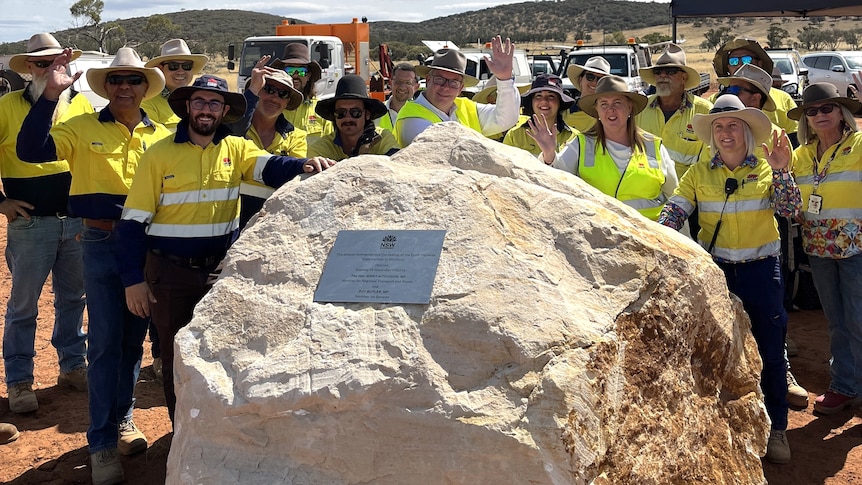 A group of men and women wearing hats and fluro clothes standing next to a big rock with a plaque on it.