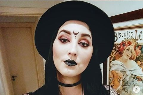 A tattoed woman in black clothing and a black hat, dark eyeliner and black lipstick, with a moon drawn between her eyebrows