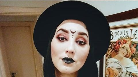 A tattoed woman in black clothing and a black hat, dark eyeliner and black lipstick, with a moon drawn between her eyebrows