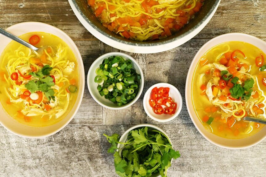 Two bowls of chicken noodle soup on a table with bowls of chilli, spring onion and corianders illustrating our simple recipe
