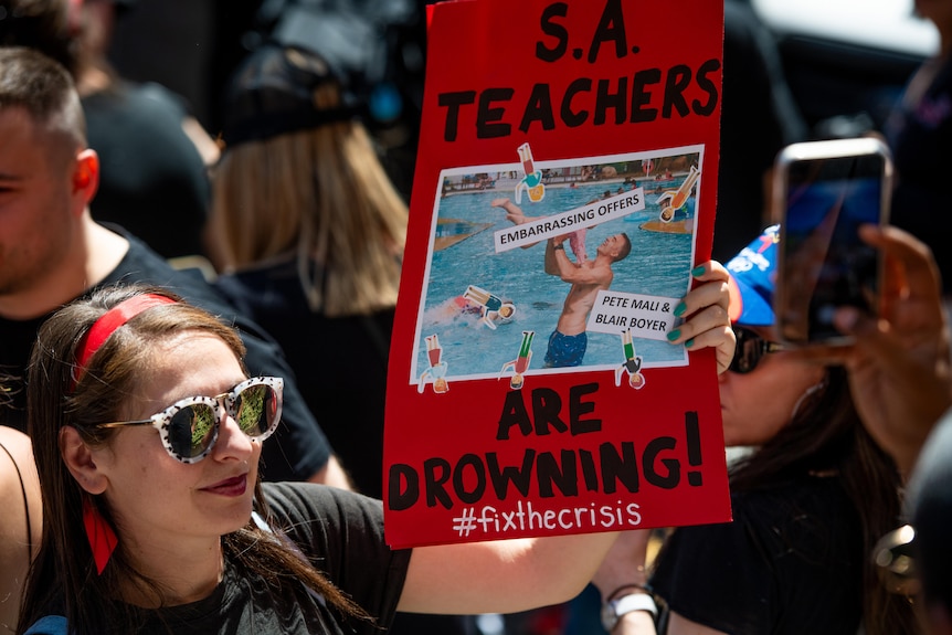 A woman wearing sunglasses and a red headband holds a red sign with a picture of Peter Malinauskas