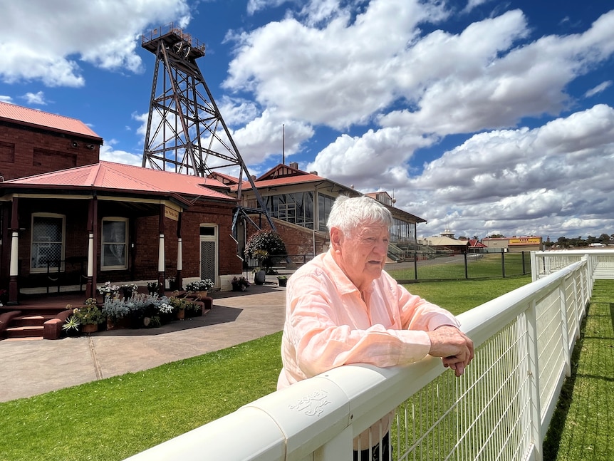 A grey haired man wearing a pink shirt stands outside the front of a country racing club