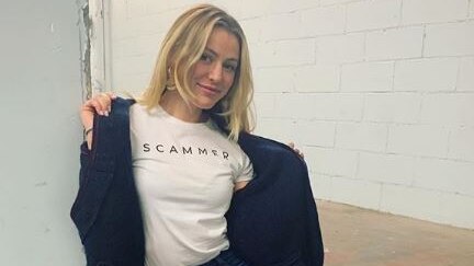 Caroline Calloway sits in a corridor wearing a t-shirt that reads "scammer", which she is holding a cardigan open to show.