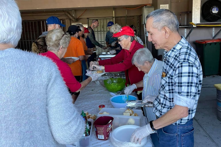 Older residents stand serving food outside at a table.