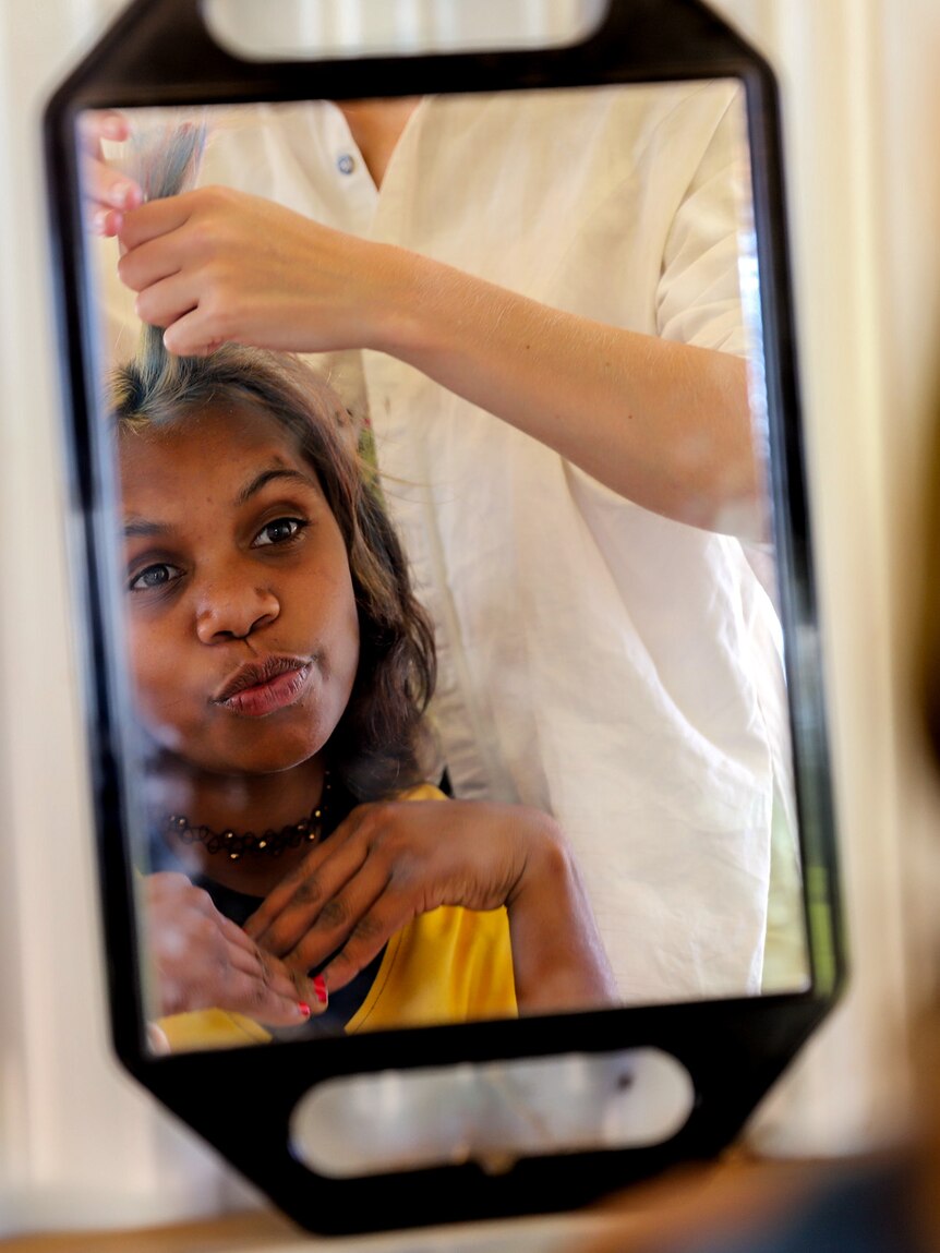 Young Aboriginal girl smiles in a mirror as she has her hair styled in a tin shed