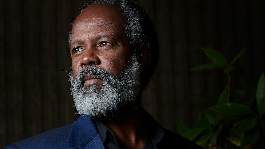 A  black man in a blue jacket with a grey beard looks off camera pensively in a tastefully lit portrait.