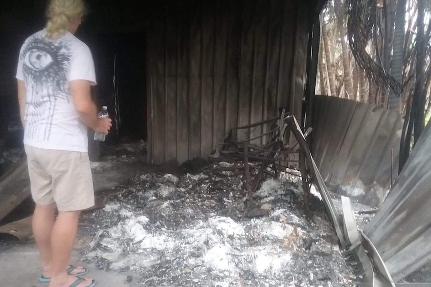 Back of man, holding water bottle, as he observes what is left of his burnt house.