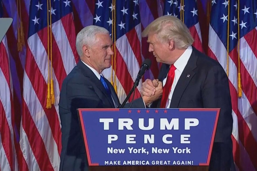Donald Trump grips Mike Pence's hand. They stand behind a lectern and in front of a row of US flags.
