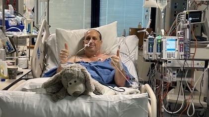 A man in a hospital bed with thumbs up and cors and machines surrounding him, with a toy dog at the foot of the bed