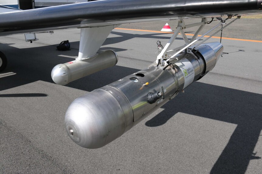 A silver canister hanging from the wing of a plane.