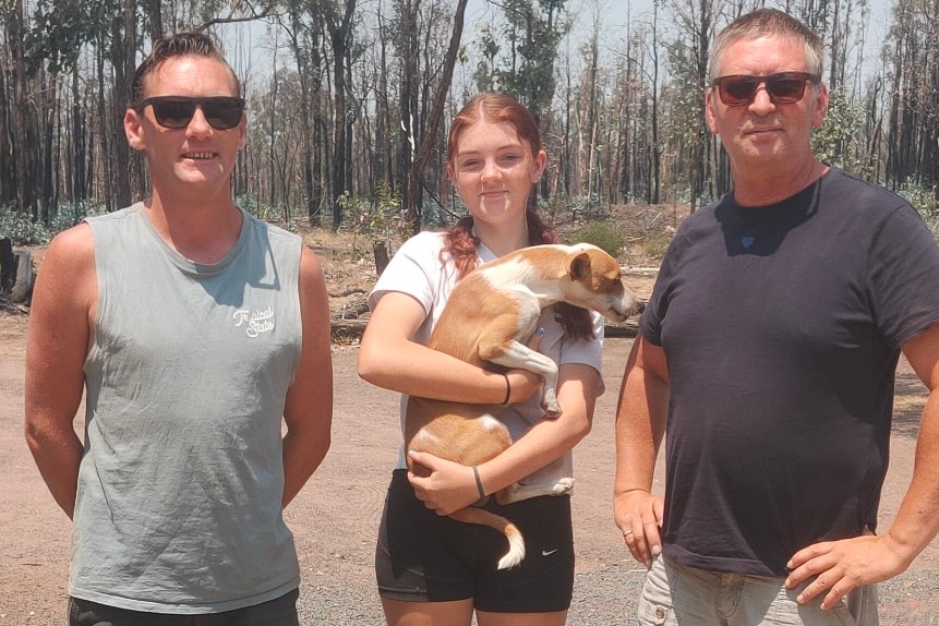 Three people, and a dog, stand in front of charred bush