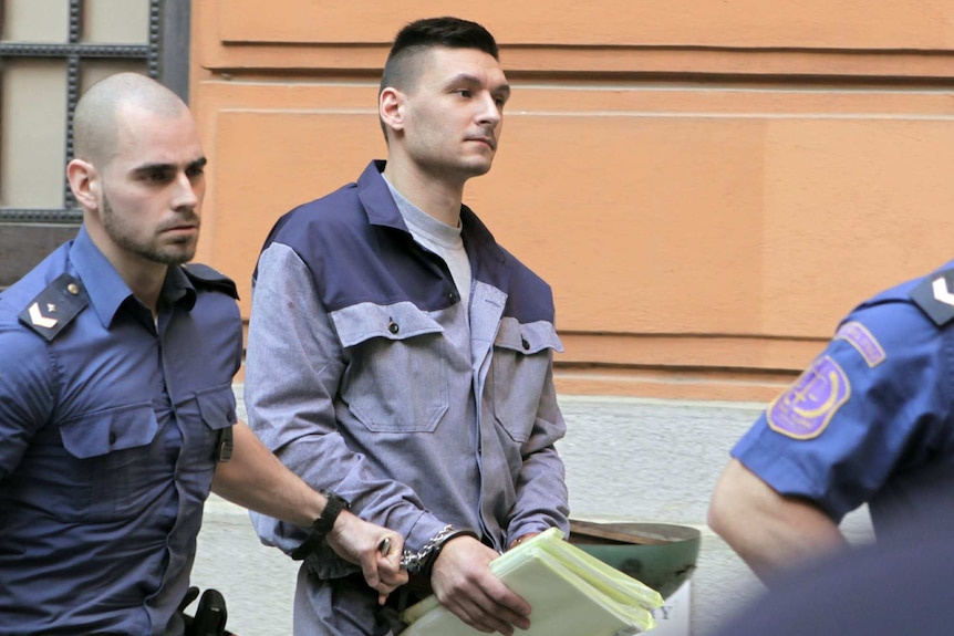 A man in shackles is escorted by security guards as he carries papers while leaving a courthouse.