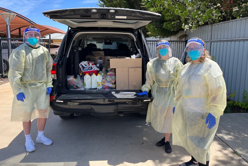 Three people dressed in full protective equipment stand at the back of a four wheel drive that's loaded with groceries