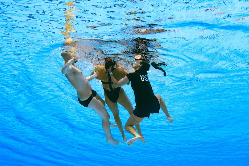 An underwater view of two people holding up an unconscious swimmer at the surface of a pool.