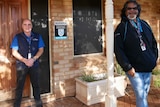 Two men stand about 1.5 metres apart smiling in front of a brick building.