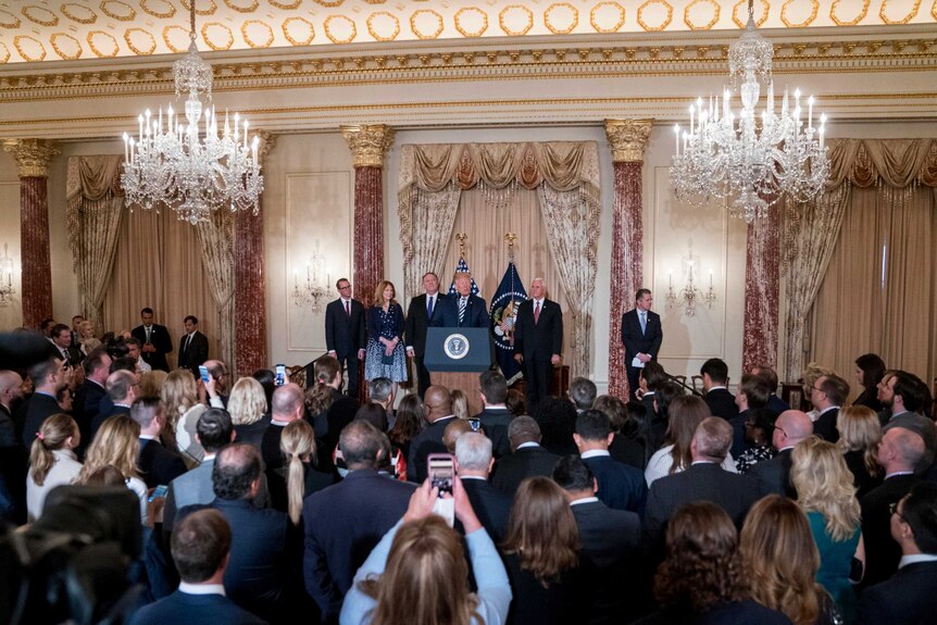 A room with chandeliers and columns filled with a crowd watching Trump speak next to Pompeo