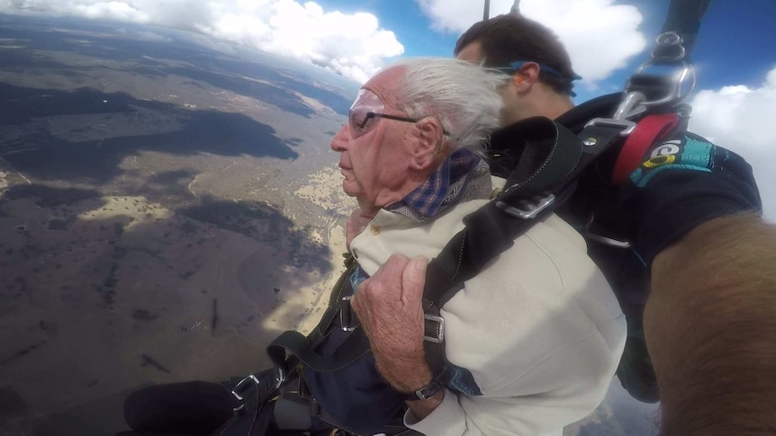 Sam clings to his parachute harness as his face and hair is buffeted by winds while he and his skydiving partner skydive.