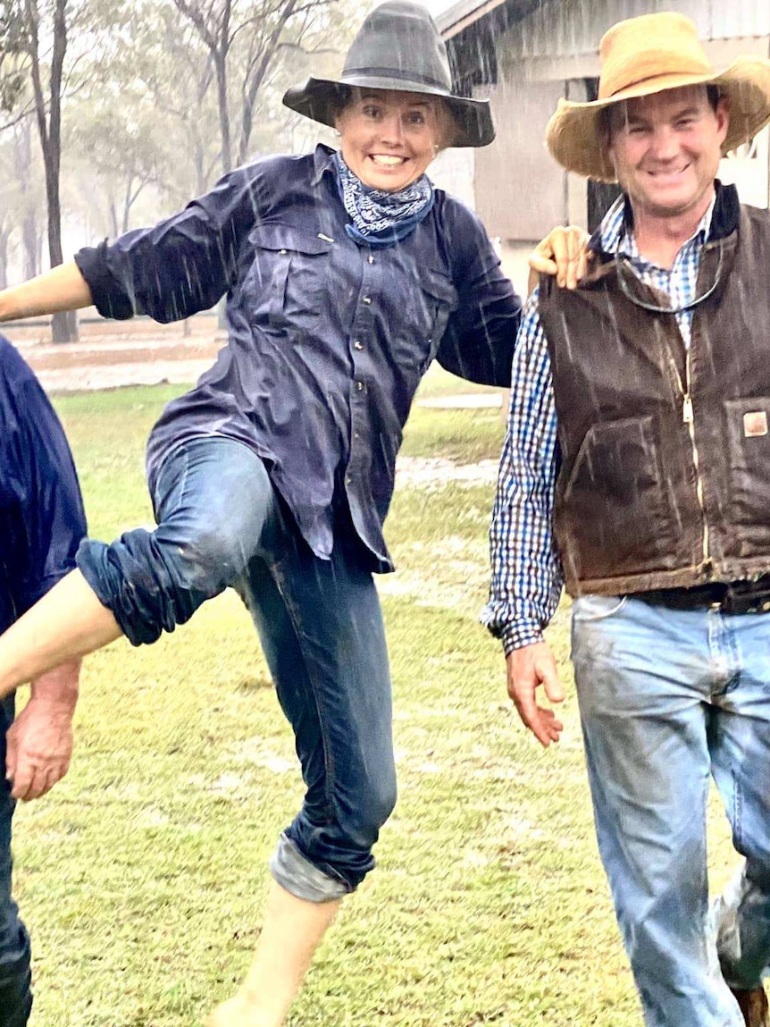 Two men and a woman walk past a farm shed in the rain, the woman jumping for joy.