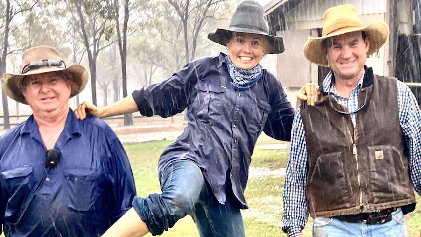 Two men and a woman walk past a farm shed in the rain, the woman jumping for joy.