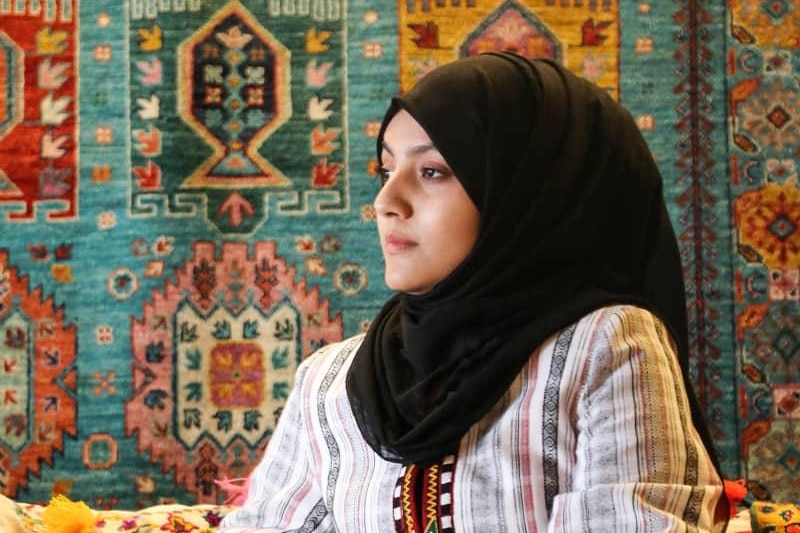 A woman wearing a headscarf sits in front of a woven rug
