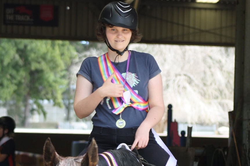 A woman wearing a helmet on horseback giving a thumbs up with a medal around her neck.