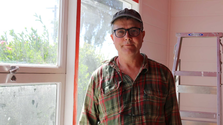 A man in a cap, glasses, and checked shirt stands in a sunny room with a ladder behind him.