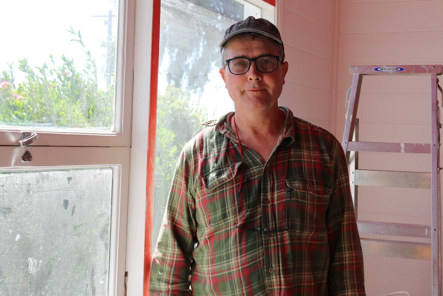 A man in a cap, glasses, and checked shirt stands in a sunny room with a ladder behind him.