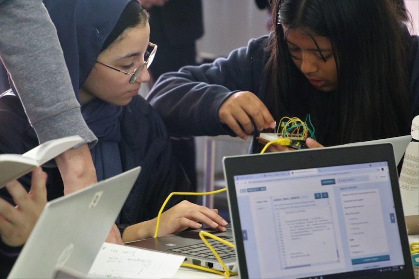 Two girls working in a school classroom, holding wires and working on computers with a teacher's arm on the desk.