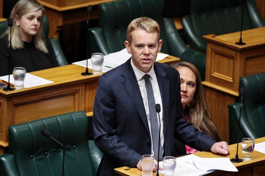 Chris Hipkins stands in parliament with two women sitting on green-leather chairs in the background