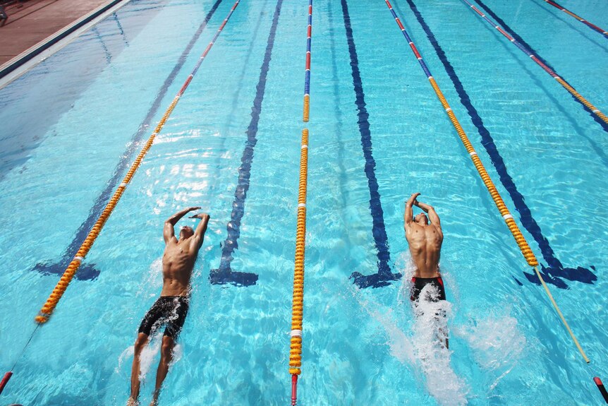 Two male swimmers, in lycra shorts, chests facing upward, push off from the end of a swimming pool, in separate lanes.