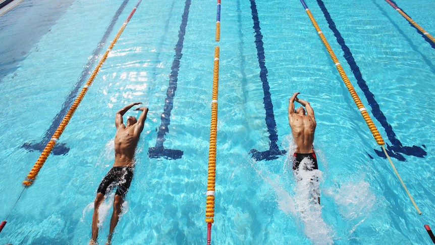 Two male swimmers, in lycra shorts, chests facing upward, push off from the end of a swimming pool, in separate lanes.