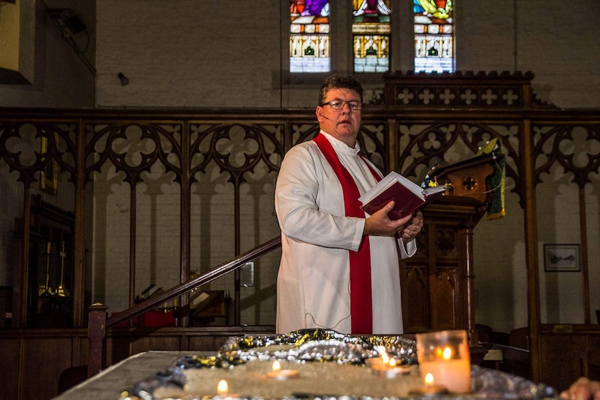 An Anglican minister in robes in a church with stained glass window in the background looks down on a small lit candle