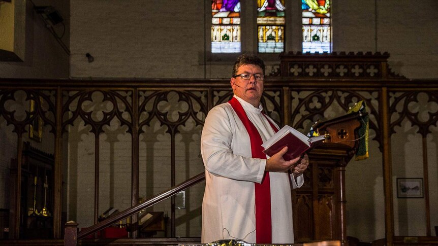 An Anglican minister in robes in a church with stained glass window in the background looks down on a small lit candle