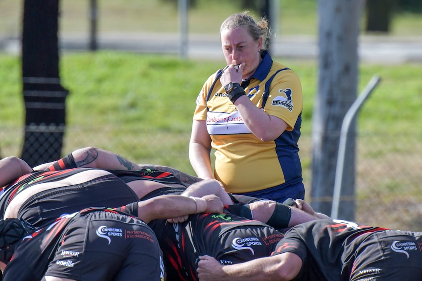 Annie blows a whistle, standing alongside a rugby scrum in her jersey.