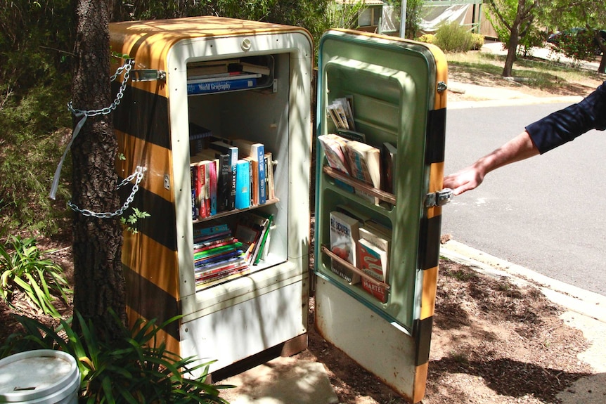 A person holds open the door of a bar fridge, which has been converted to house books.