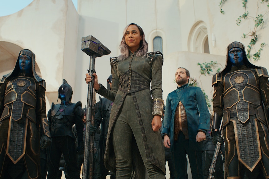 A black woman with pastel pink hair wearing a fierce grey battle suit stands in front of an amry of statuesque soldiers.