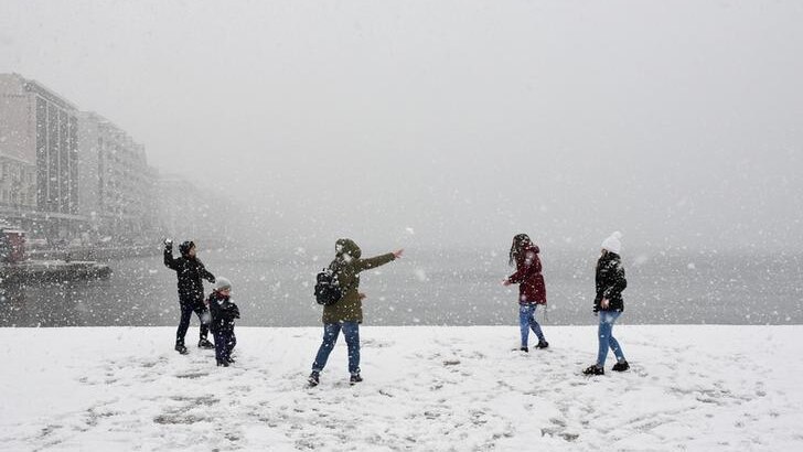 A group of five people launch snowballs near the sea, with buildings to their left during heavy snowfall.