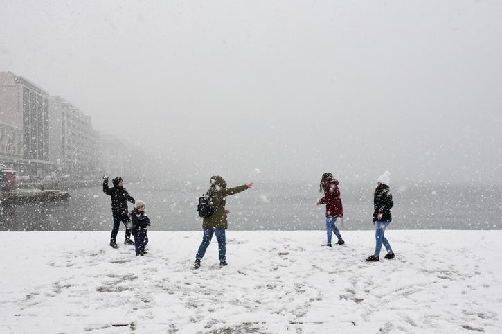 A group of five people launch snowballs near the sea, with buildings to their left during heavy snowfall.